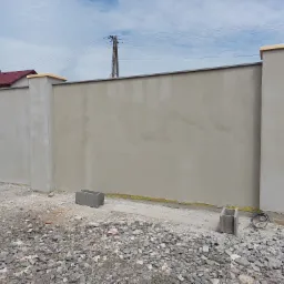 Świeżo otynkowany fragment muru oporowego z filarami, widoczne bloczki betonowe i kabel elektryczny na żwirowym podłożu.