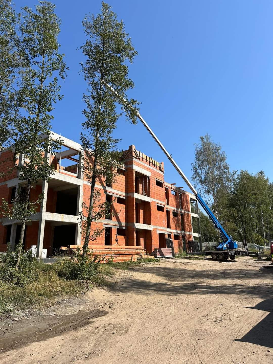Wznoszenie trzypiętrowego budynku z czerwonej cegły ceramicznej, widoczny dźwig teleskopowy i ułożone deski, teren budowy w słoneczny dzień.