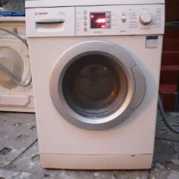 Biała pralka Bosch Maxx 7 z wyświetlaczem pokazującym '0:09', stojąca na betonowej posadzce, z widoczną inną pralką w tle.