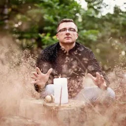 Mężczyzna w okularach medytuje w lesie przed płonącą świecą, unosząc dłonie w geście skupienia.