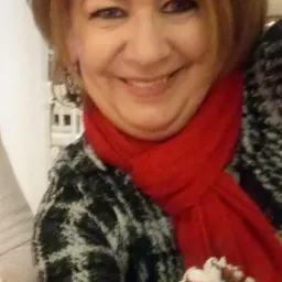 Uśmiechnięta kobieta z rudymi włosami i czerwonym szalem, trzymająca deser lodowy z bitą śmietaną i sosem czekoladowym w szklanym pucharku.