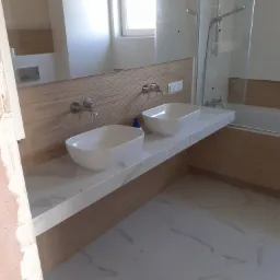 Dwie białe, owalne umywalki nablatowe z chromowanymi bateriami ściennymi na blacie z białego marmuru, drewnopodobna okładzina ściany, kabina prysznicowa z transparentnego szkła, białe płytki...