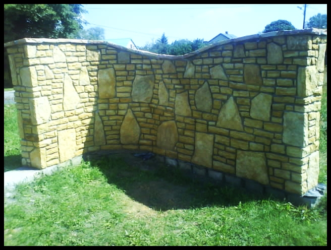 Ogrodzenie z piaskowca w ogrodzie, zbudowane z nieregularnych bloków i kamieni o ciepłej, żółto-beżowej barwie. Fragment trawnika widoczny u podstawy.