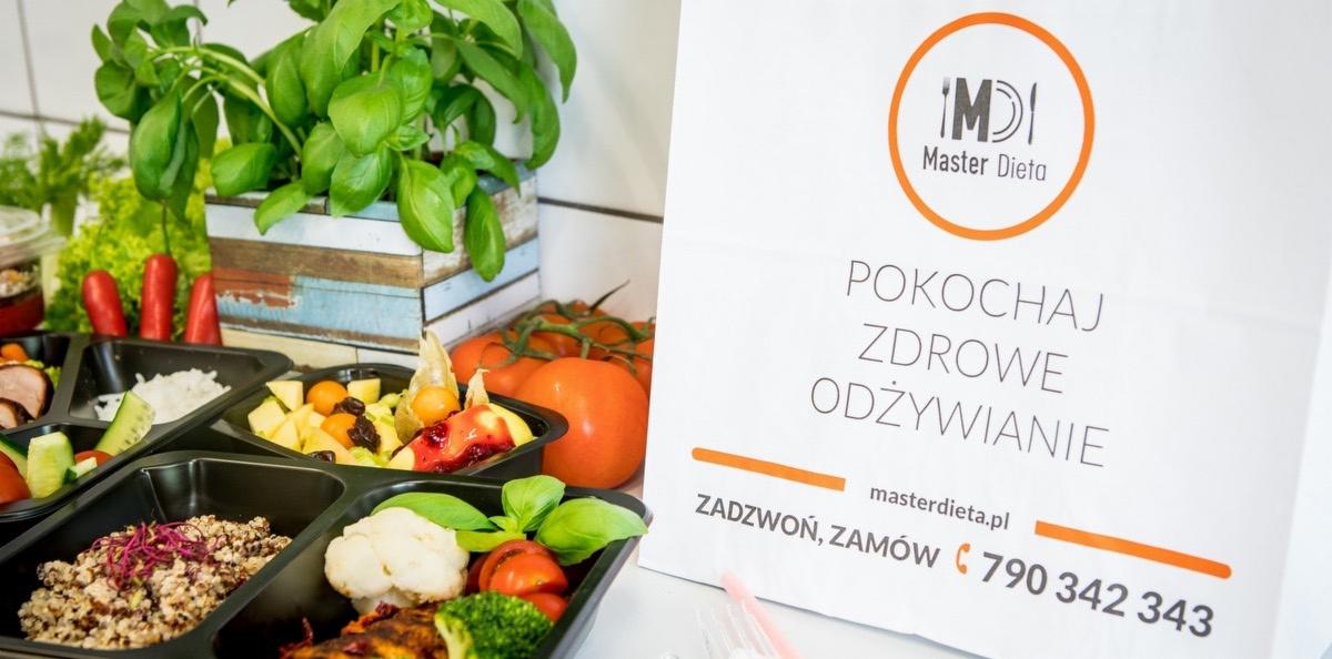 Zestaw posiłków dietetycznych w czarnych pojemnikach, zróżnicowane dania, obok torba z logo firmy cateringowej, świeże zioła i pomidory w tle.