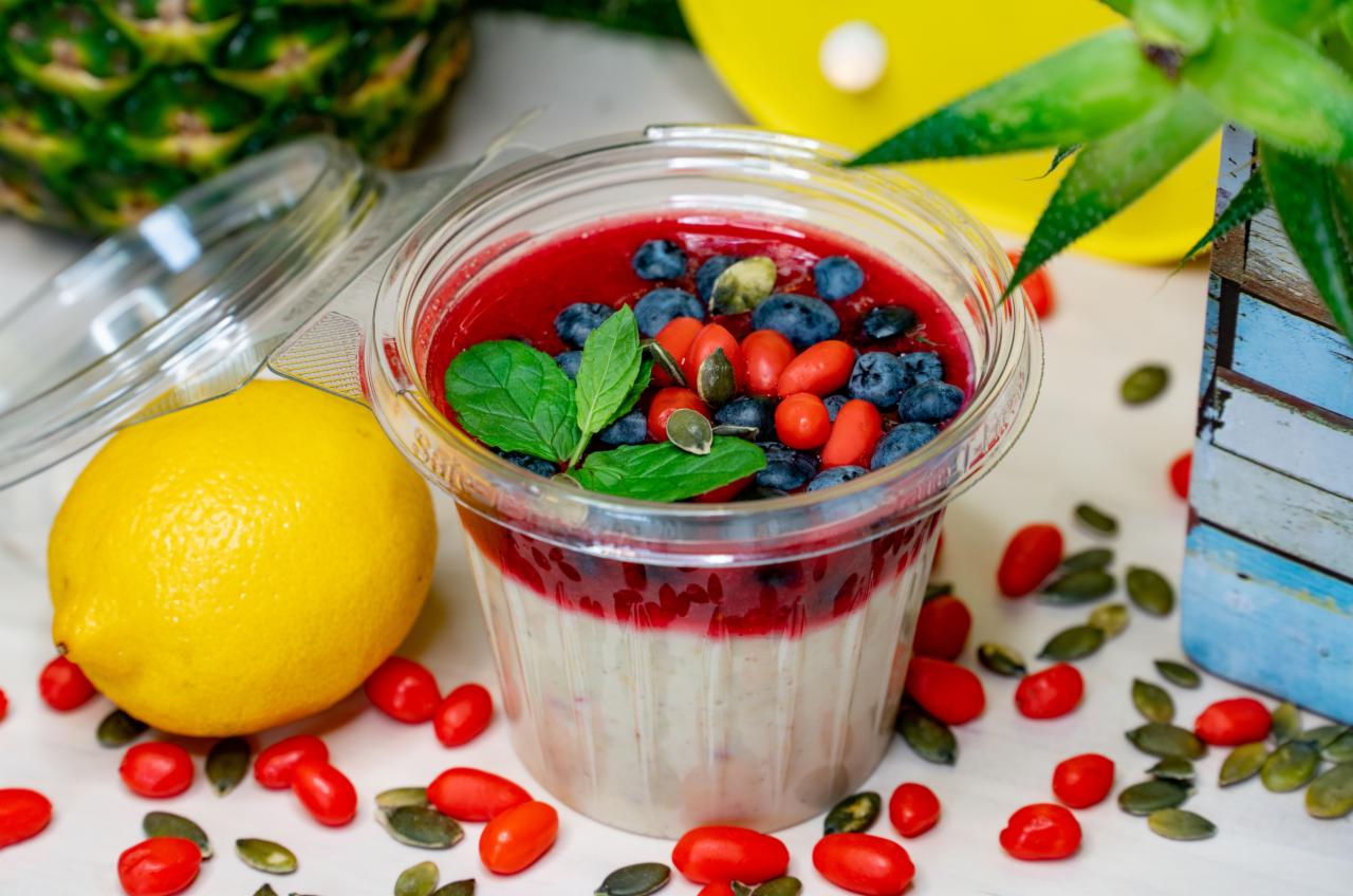 Deser dietetyczny w przezroczystym pojemniku z warstwami białej masy, czerwonego musu i świeżych borówek, goji oraz listkami mięty, obok cytryna i ananas w tle.