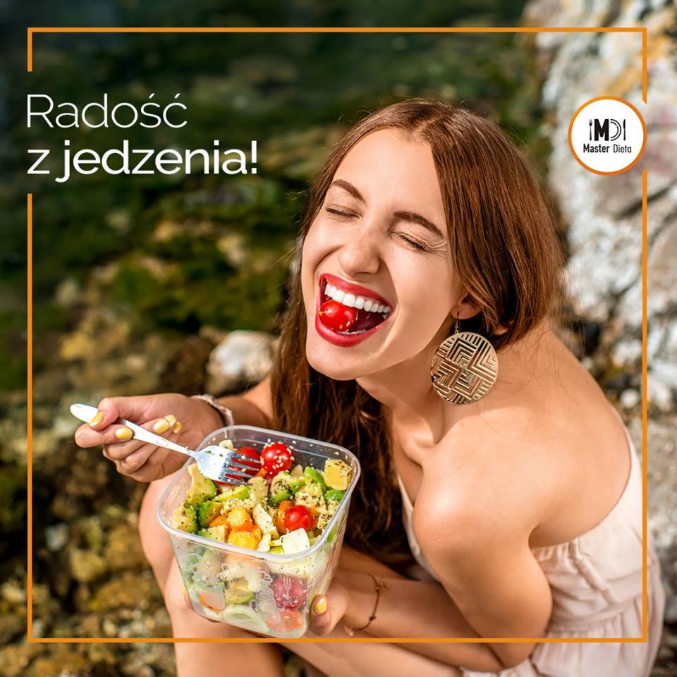 Uśmiechnięta kobieta z pomidorkiem w ustach, trzymająca pojemnik z sałatką warzywną i widelec, na tle naturalnego otoczenia i logo firmy.