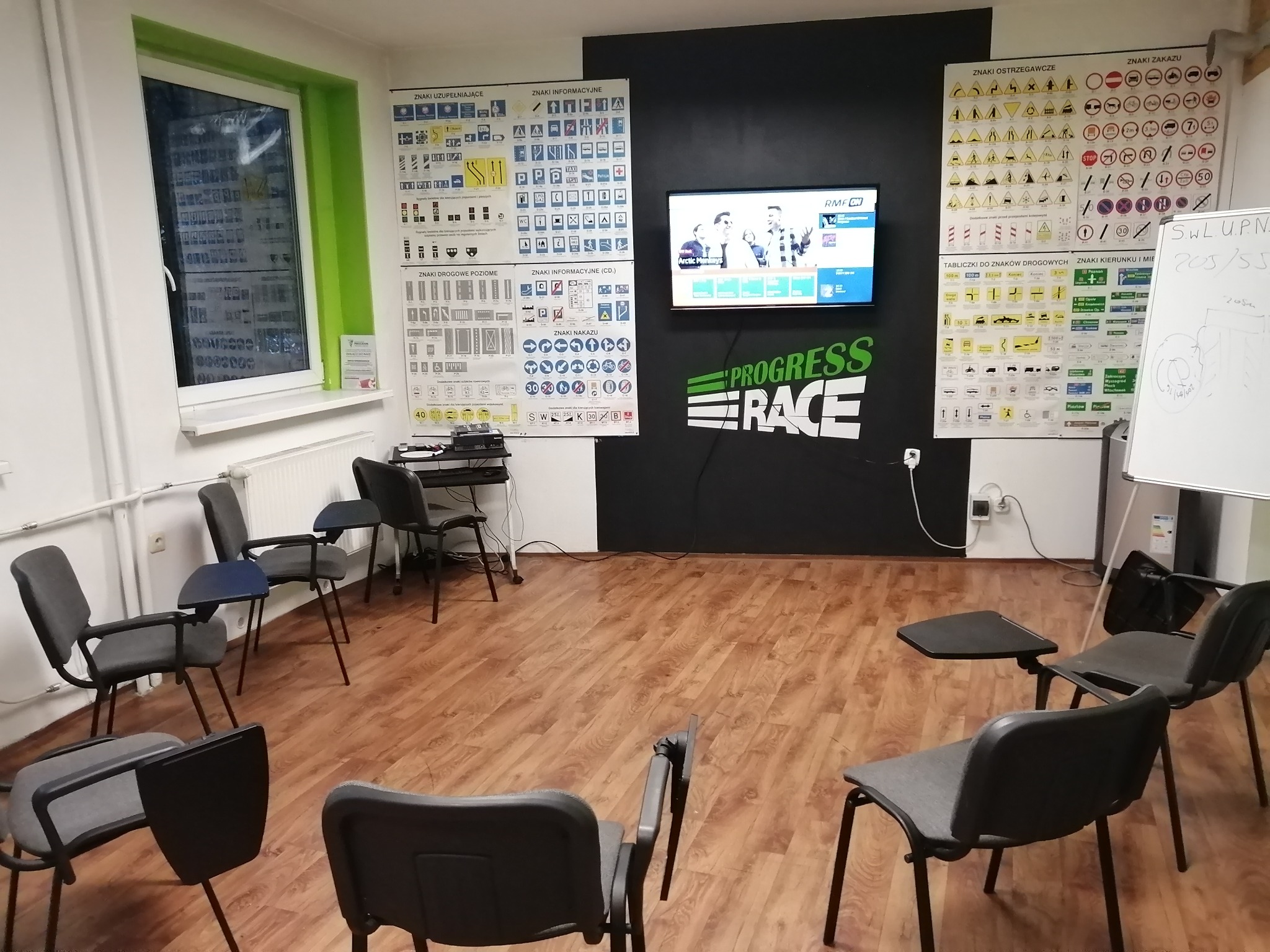 Sala wykładowa z krzesłami z pulpitem, tablicami ze znakami drogowymi i telewizorem wyświetlającym scenę z filmu, logo 'Progress Race' na ścianie.