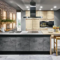 Nowoczesna kuchnia z szarą wyspą i blatem z jasnego drewna, ceglana ściana z półkami na zioła, stalowy okap i drewniane szafki w tle, oświetlenie punktowe.