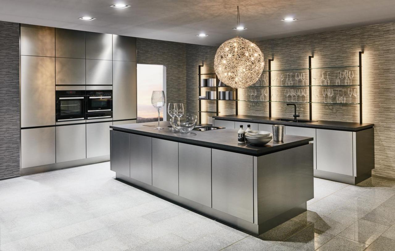 Nowoczesna kuchnia z wyspą, stalowe fronty szafek, oświetlenie punktowe i dekoracyjna lampa wisząca w kształcie kuli. Na blacie szklane naczynia i akcesoria kuchenne.