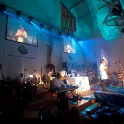 Wnętrze kościoła zaadaptowane na salę koncertową, scena z orkiestrą i chórem podświetlona niebieskim światłem, na ścianach wyświetlane projekcje w tle.