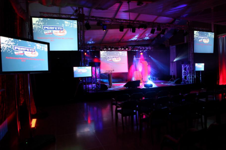 Scena z nagłośnieniem i oświetleniem przygotowana na konferencję, z licznymi ekranami wyświetlającymi logo firmy, krzesła dla publiczności, reflektory i efekty świetlne w kolorach czerwonym...