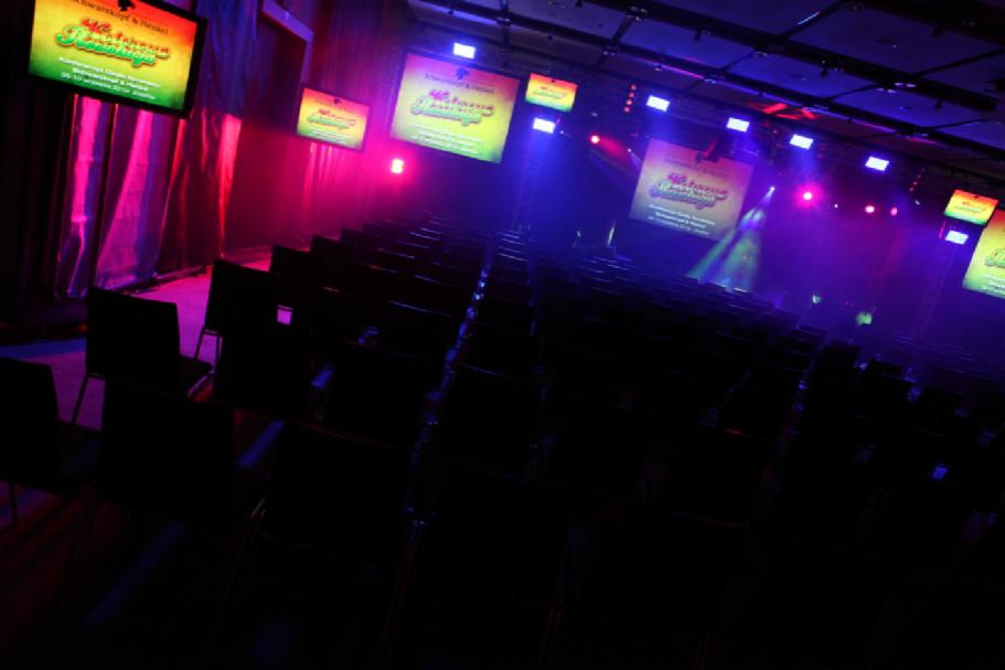 Sala konferencyjna z rzędami krzeseł, oświetlona kolorowymi światłami sceny i ekranami wyświetlającymi logo wydarzenia.