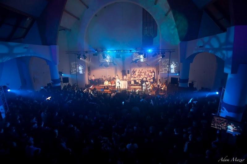 Widok z góry na koncert w dużej sali z chórem i orkiestrą na scenie oświetlonej niebieskim światłem, z publicznością w ciemności.