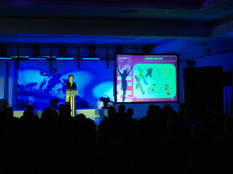 Prelegentka na scenie podczas prezentacji z projektorem wyświetlającym slajd z grafiką, oświetlenie sceniczne w kolorze niebieskim, widownia w tle.