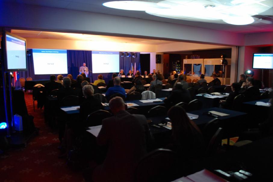 Konferencja w sali hotelowej: prelegent na scenie, publiczność siedząca przy stolikach z dokumentami, ekrany projekcyjne z prezentacją, oświetlenie sceniczne.