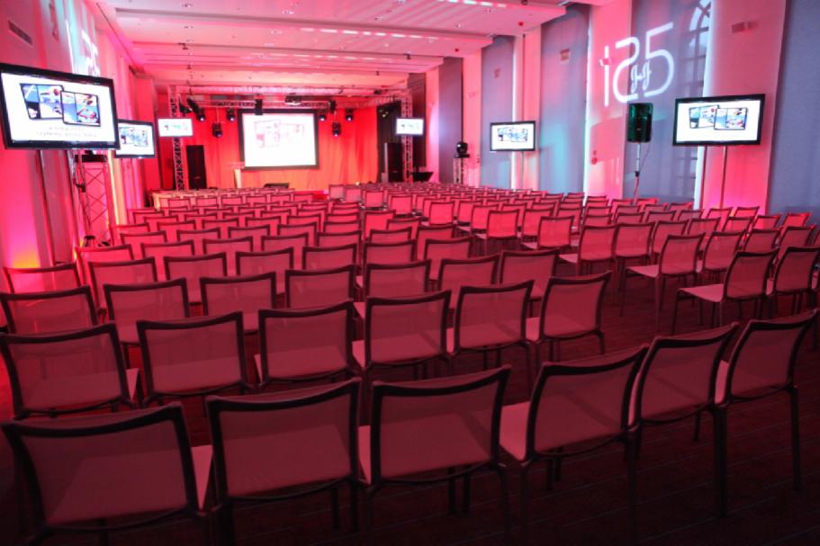 Sala konferencyjna z rzędami krzeseł, sceną z ekranem, oświetleniem w odcieniach czerwieni i ekranami LCD po bokach, przygotowana na wydarzenie.