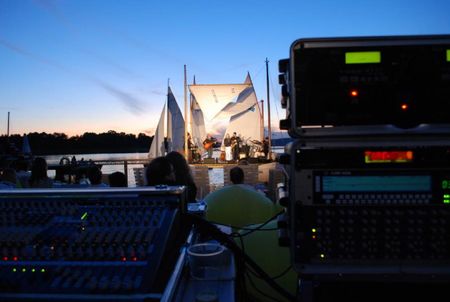 Koncert na wodzie o zmierzchu, scena zbudowana na platformie z żaglami, widok zza konsolety nagłośnieniowej, w tle publiczność.