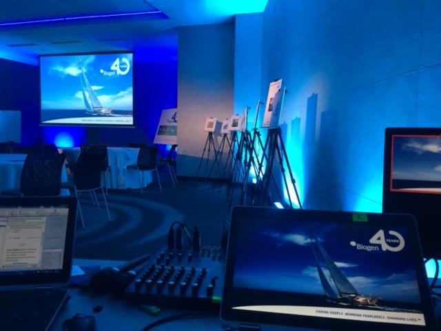 Sala konferencyjna w niebieskim oświetleniu z ekranem projekcyjnym wyświetlającym żaglówkę i logo 'Biogen 40 Years', mikser dźwięku i laptopy na pierwszym planie, plakaty na sztalugach w tle.