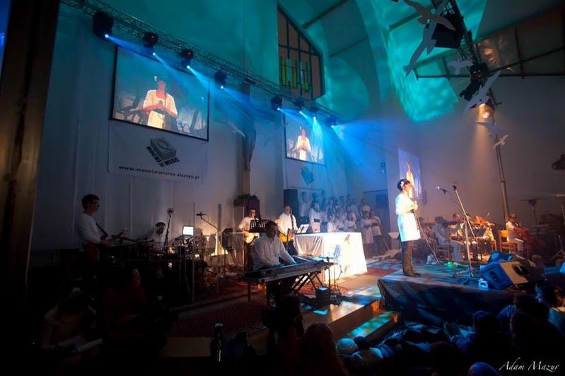 Wnętrze kościoła zaadaptowane na salę koncertową, scena z orkiestrą i chórem podświetlona niebieskim światłem, na ścianach wyświetlane projekcje w tle.