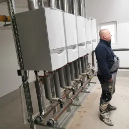 Instalacja rzędu pieców gazowych Vaillant z odprowadzeniem spalin, rurami i armaturą, nadzorowana przez pracownika w kombinezonie roboczym, widok z boku.