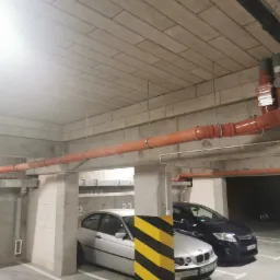 Pomarańczowe rury instalacji przeciwpożarowej pod sufitem parkingu podziemnego, widoczne samochody i filar z czarno-żółtymi pasami.