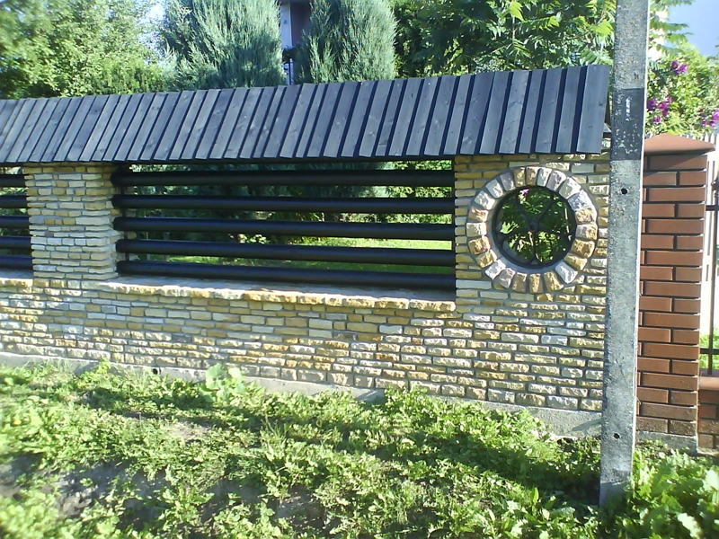 Fragment ogrodzenia z piaskowca z drewnianym zadaszeniem i okrągłym oknem z kamienia, widoczne czarne rury poziome oraz słup elektryczny w tle.