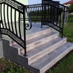 Zewnętrzne schody wejściowe wyłożone płytkami imitującymi drewno, z czarną metalową balustradą zdobioną motywem greckim.