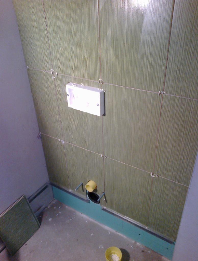 Układanie pionowych, zielonych płytek z fakturą na ścianie w łazience, widoczne krzyżyki dystansowe, puszka elektryczna i fragment instalacji wodnej.