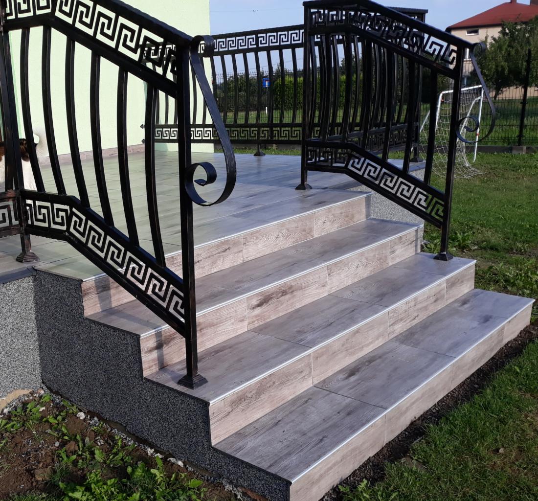 Zewnętrzne schody wejściowe wyłożone płytkami imitującymi drewno, z czarną metalową balustradą zdobioną motywem greckim.