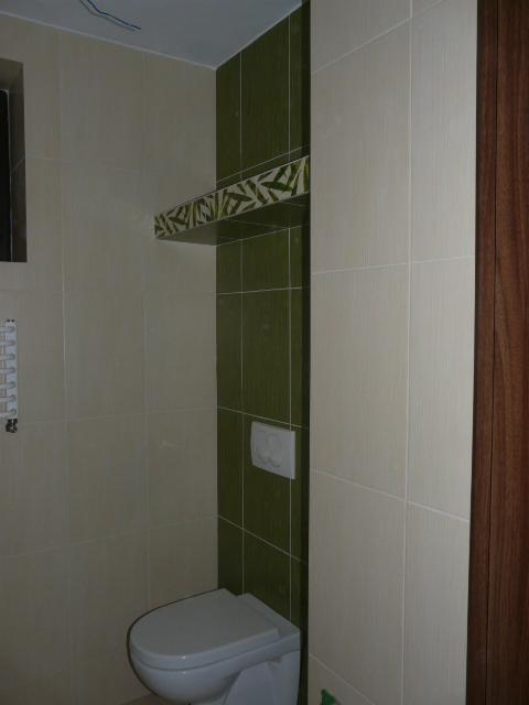 Wykończona łazienka z kremowymi i zielonymi płytkami, wisząca półka z motywem roślinnym nad toaletą, widoczne pionowe płytki w odcieniach zieleni.