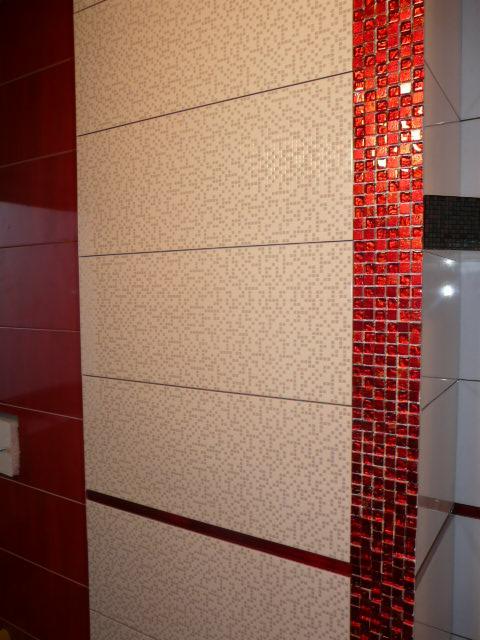 Ściana wyłożona beżowymi płytkami z drobnym wzorem, z pionowym pasem dekoracyjnej, błyszczącej, czerwonej mozaiki.