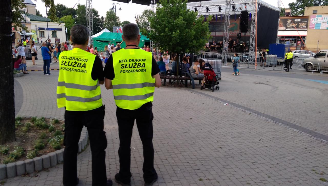Dwaj mężczyźni w żółtych kamizelkach odblaskowych z napisem 'RED-DRAGON SŁUŻBA PORZĄDKOWA' stoją tyłem do kamery na tle plenerowej sceny z zespołem muzycznym podczas imprezy miejskiej.