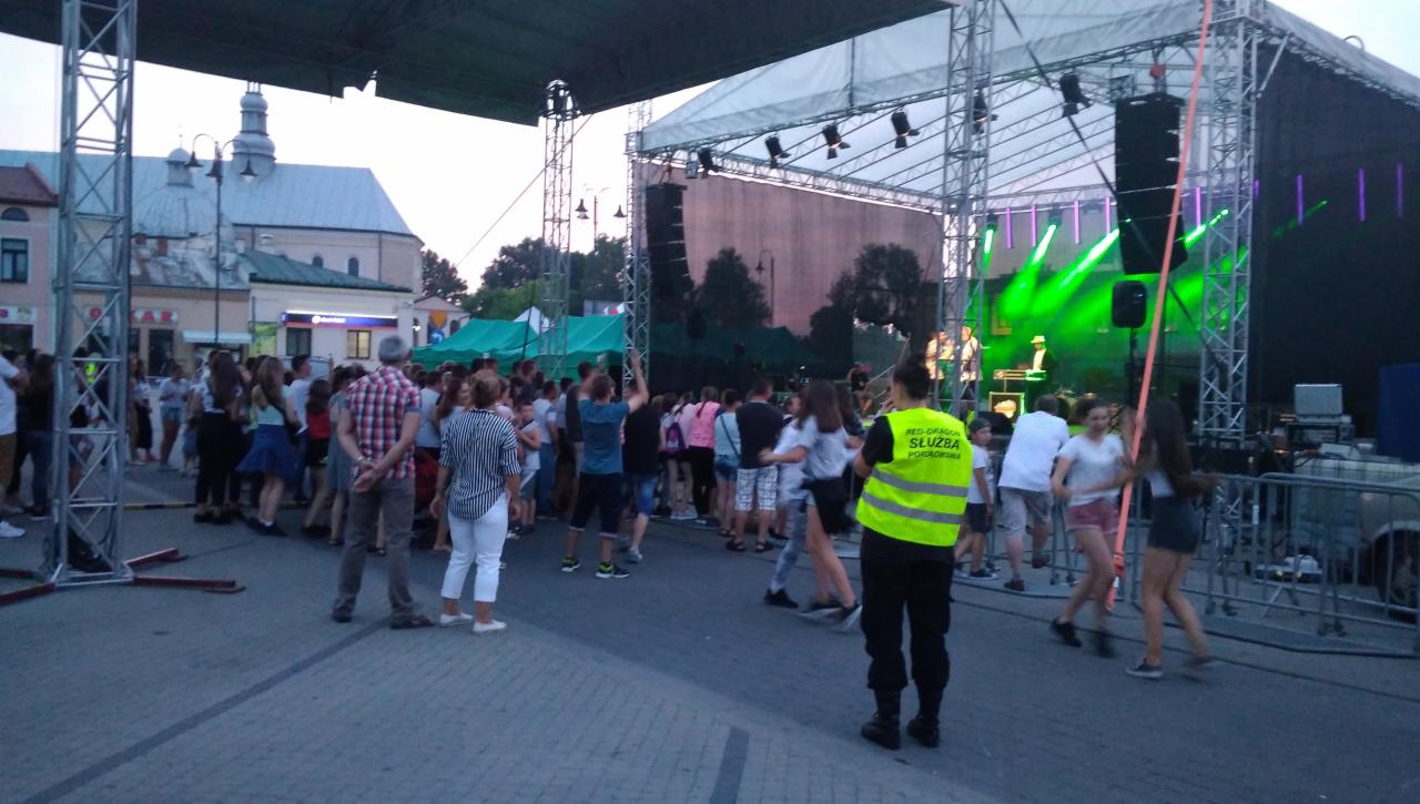 Ochrona imprezy masowej: Funkcjonariusz w kamizelce odblaskowej stoi tyłem do kamery, kontrolując tłum przed sceną oświetloną zielonymi laserami podczas koncertu plenerowego w tle widać budynek...