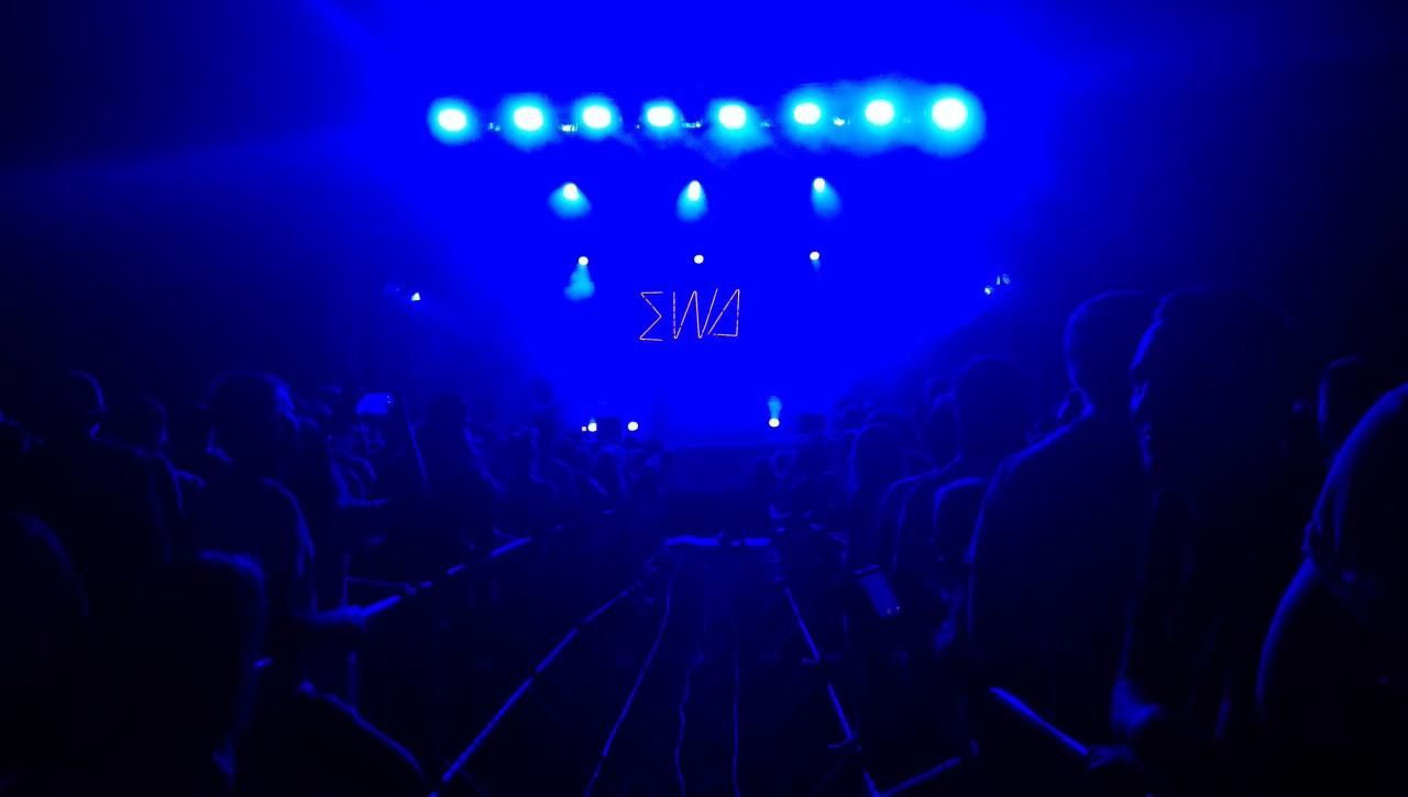 Tłum ludzi w ciemności oświetlony niebieskimi światłami, na scenie napis 'ΣWA'.