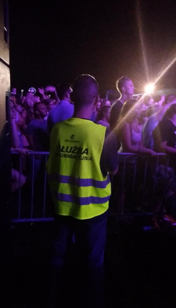 Ochroniarz w jaskrawej kamizelce z napisem 'Służba Informacyjna' stoi tyłem do kamery, obserwując tłum ludzi za metalowymi barierkami podczas nocnego wydarzenia.