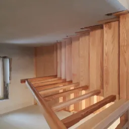 Surowe drewniane schody bez podstopnic, montowane w pomieszczeniu w trakcie wykańczania; widoczne elementy konstrukcyjne i surowy beton na ścianach.