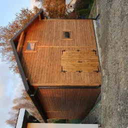 Drewniana konstrukcja z dwuspadowym dachem, małe okna i ozdobne okucia na drzwiach, widoczna w jesiennym świetle na tle drzew.