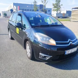 Czarny Citroen Picasso z żółtą naklejką 'Wrocław 5117' na drzwiach, zaparkowany na asfalcie przed budynkiem przemysłowym w słoneczny dzień.