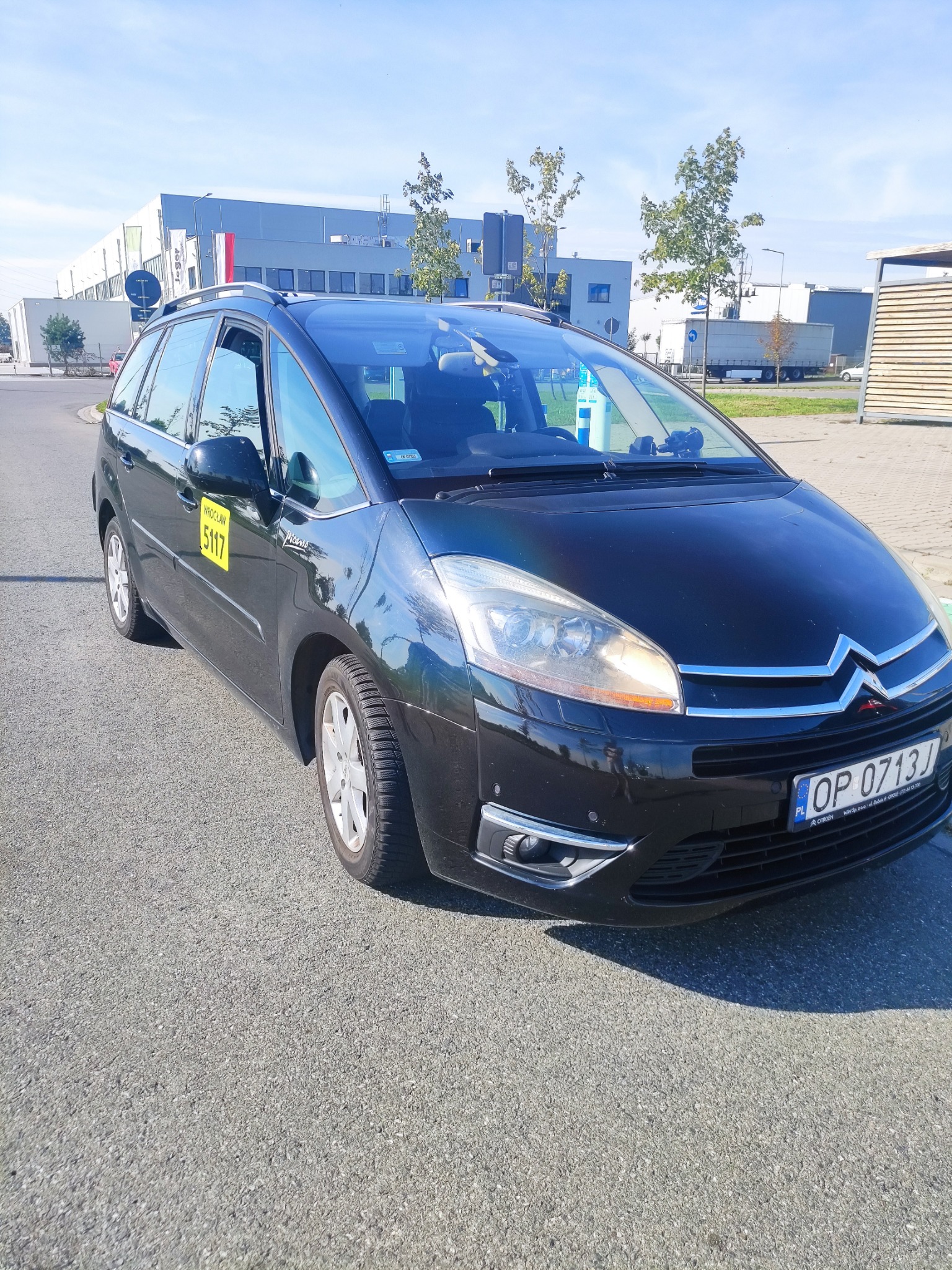Czarny Citroen Picasso z żółtą naklejką 'Wrocław 5117' na drzwiach, zaparkowany na asfalcie przed budynkiem przemysłowym w słoneczny dzień.