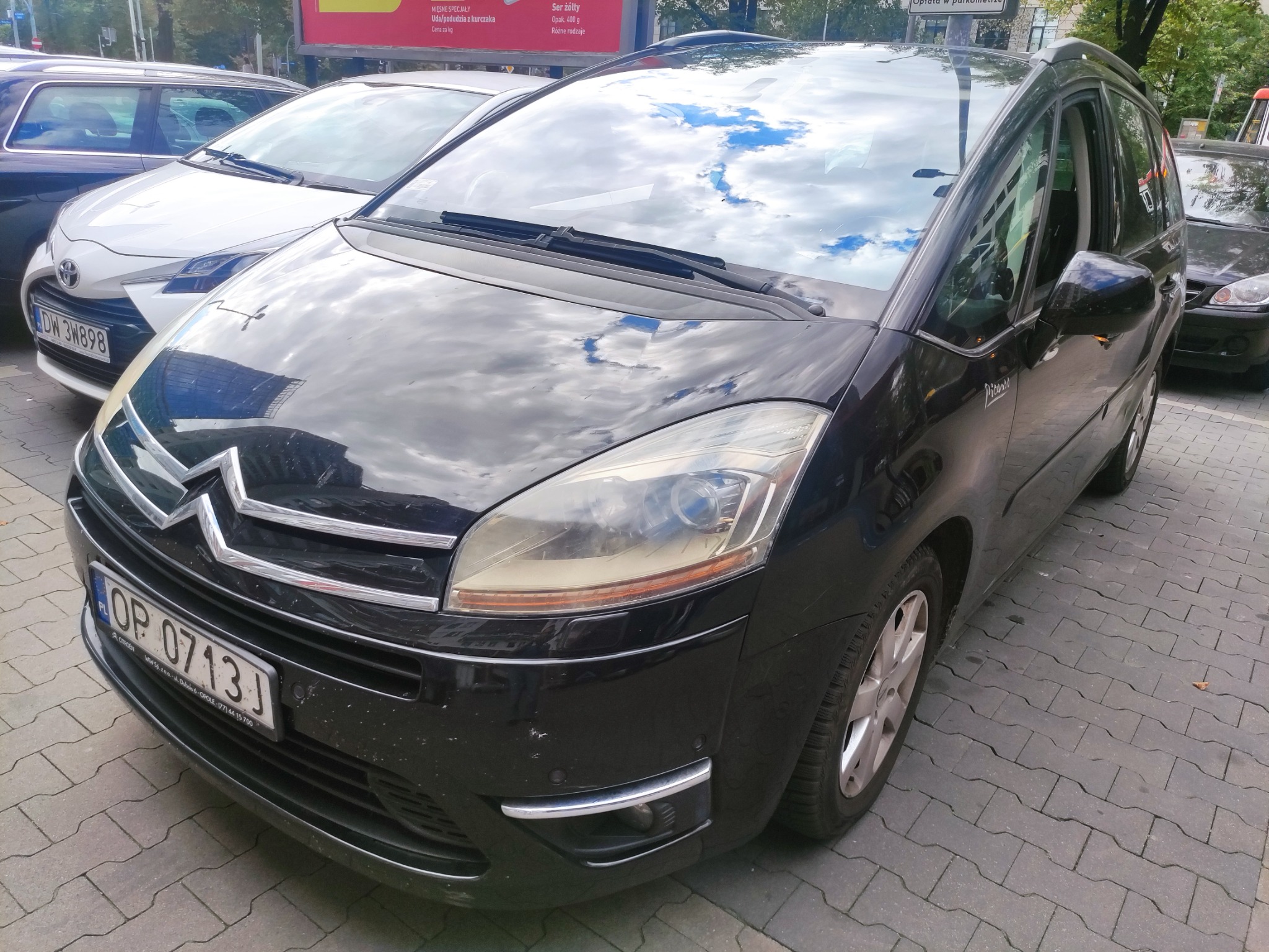 Czarny Citroen Picasso zaparkowany na chodniku z kostki brukowej, widoczne odbicie chmur w szybie, tablica rejestracyjna OP 0713J. Na drugim planie zaparkowane inne samochody oraz reklama.