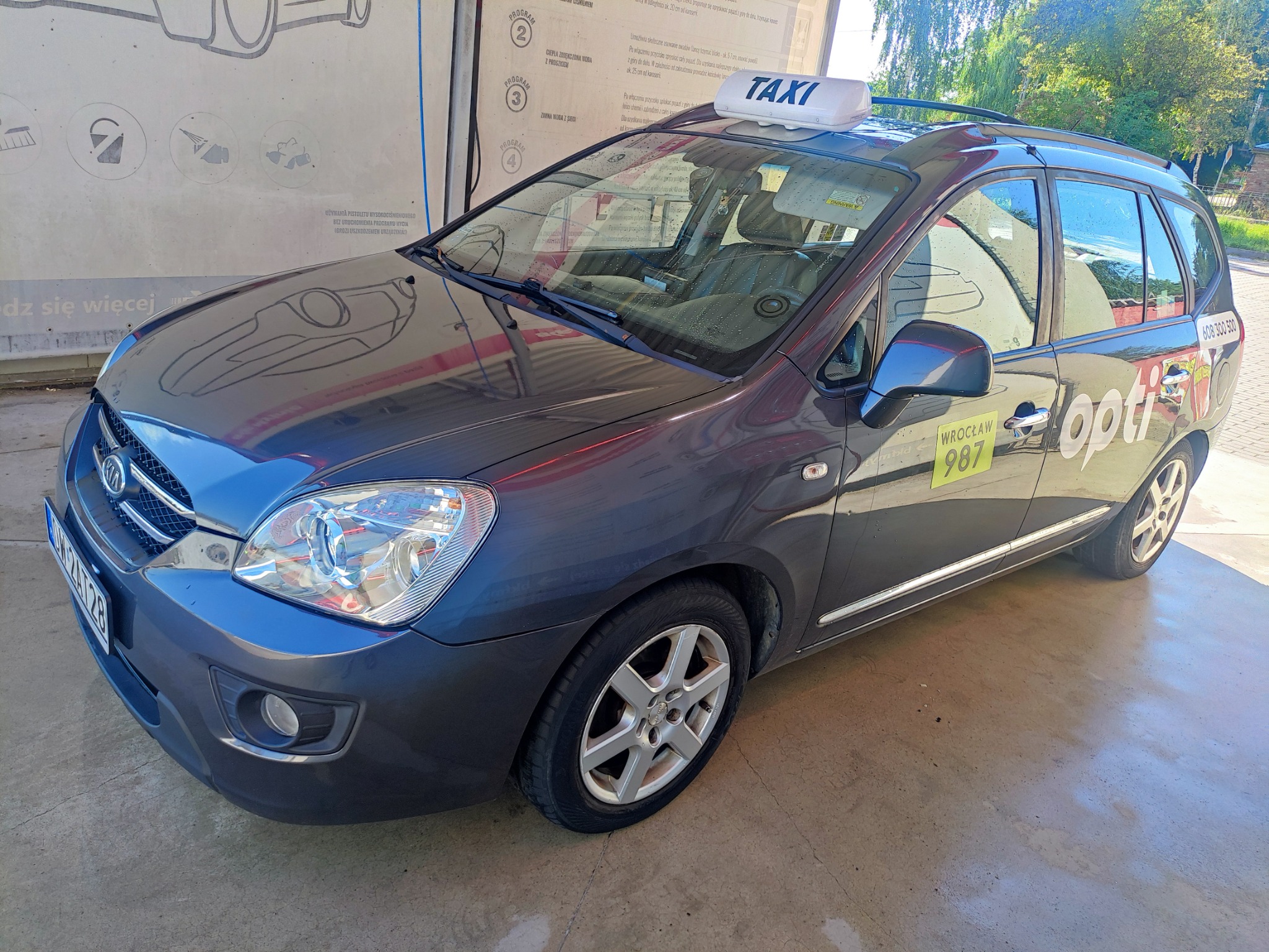 Szary samochód osobowy z napisem TAXI na dachu, zaparkowany przed myjnią samochodową, z naklejką 'opti' i numerem bocznym 987.