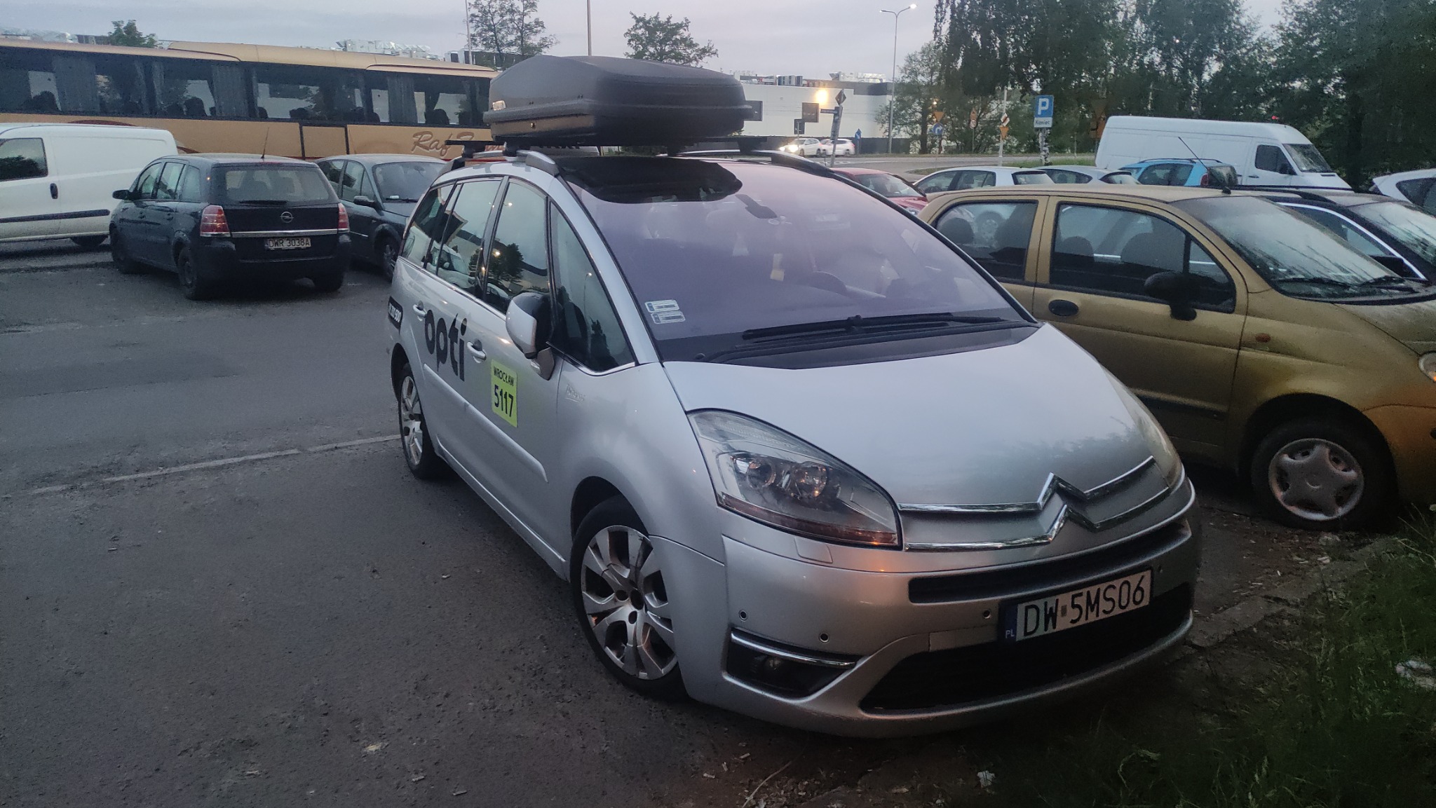 Srebrny Citroen Picasso z bagażnikiem dachowym na parkingu, z widocznym logo OPTI i numerem bocznym 5117, zaparkowany obok innych samochodów i autobusu.