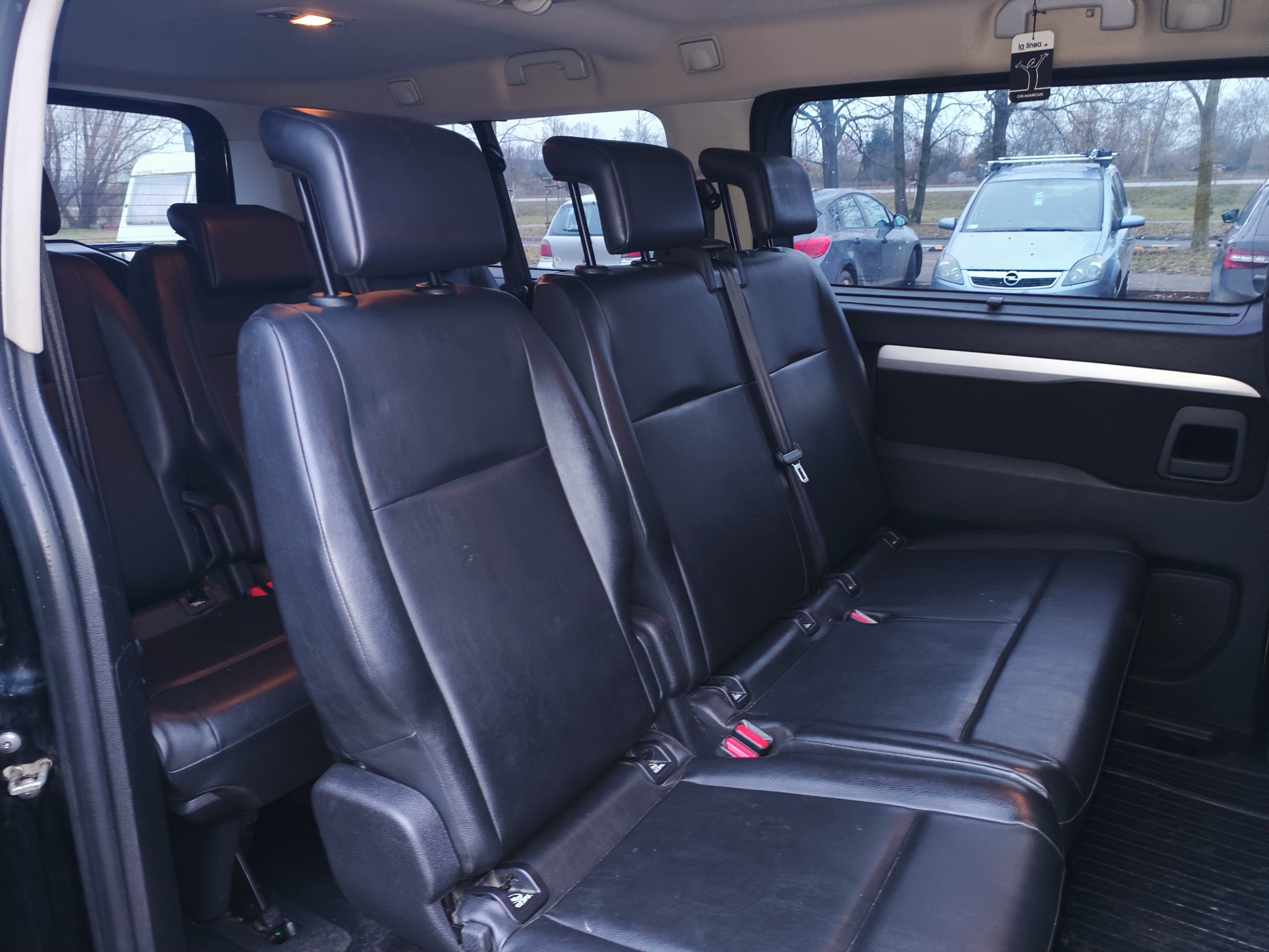 Wnętrze busa z trzema rzędami czarnych, skórzanych siedzeń. Widok z boku, z zewnątrz widać zaparkowane samochody i drzewa. Ciemna tapicerka i gumowa podłoga.