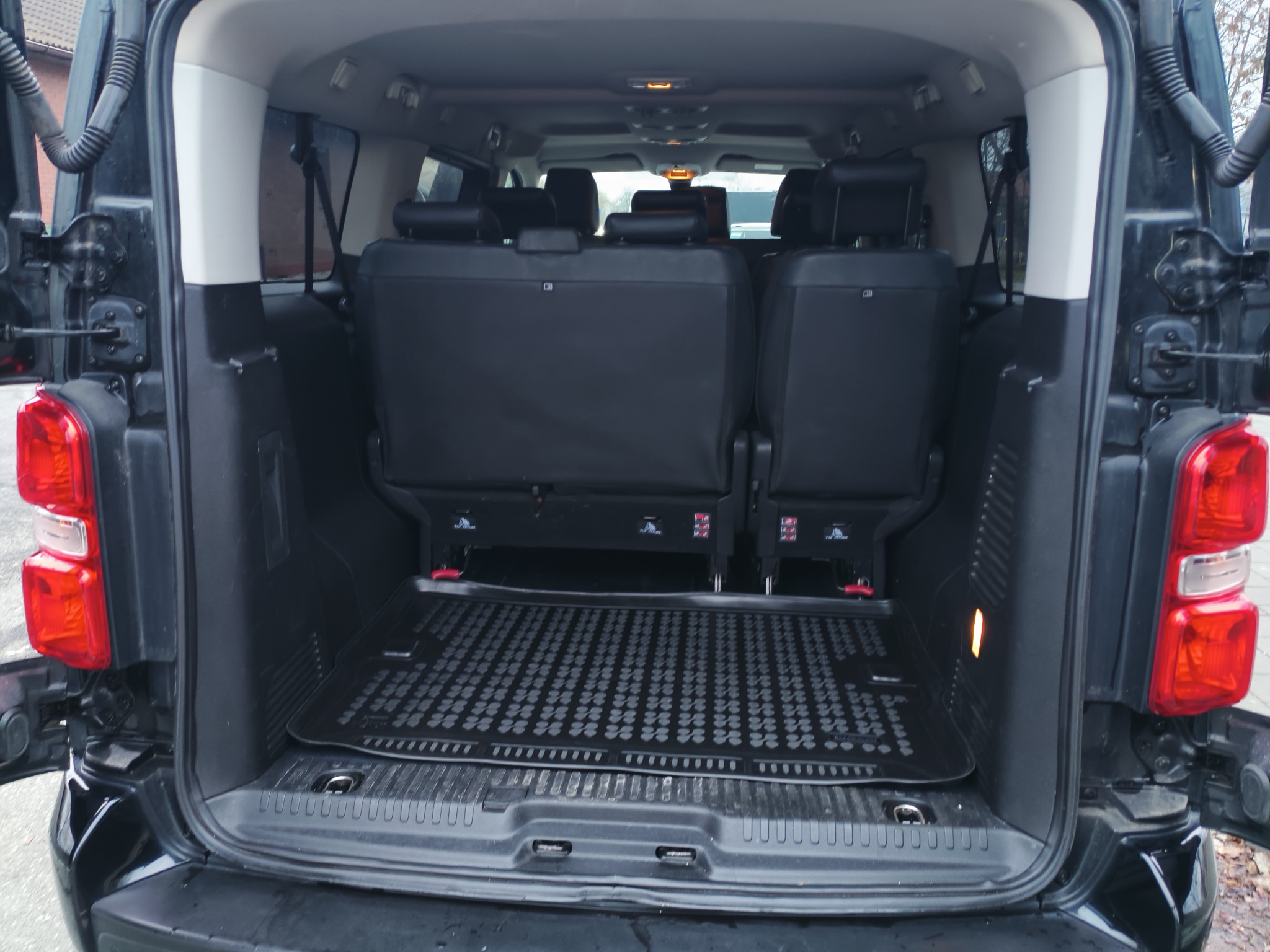 Wnętrze bagażnika czarnego busa z rozłożonymi siedzeniami i gumową wycieraczką. Widoczne otwarte drzwi bagażnika i światła stop.