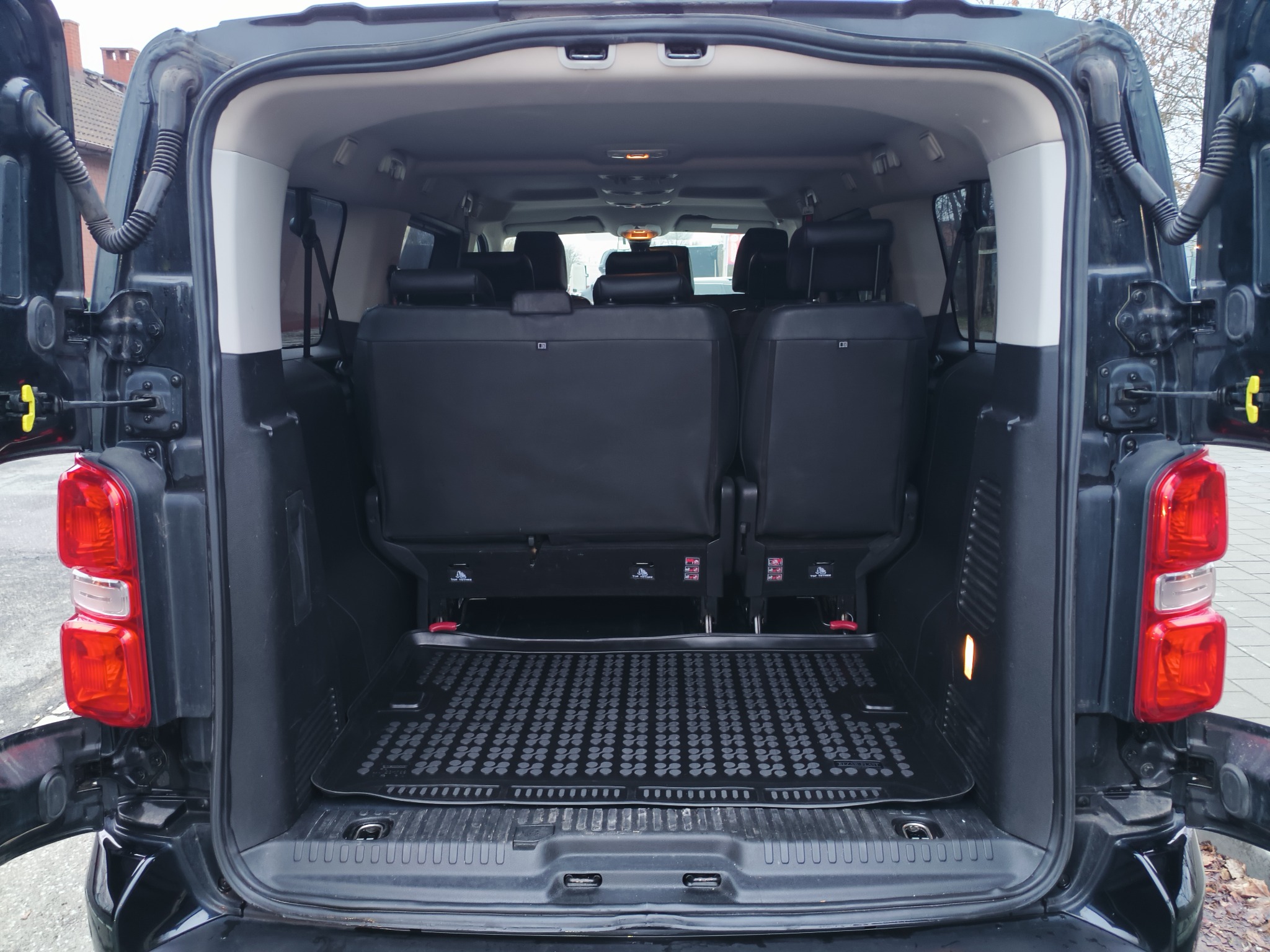 Otwarty bagażnik czarnego busa z widocznymi siedzeniami i gumową wycieraczką. Widok z zewnątrz, światło dzienne. Gotowy do załadunku bagaży lub towarów.