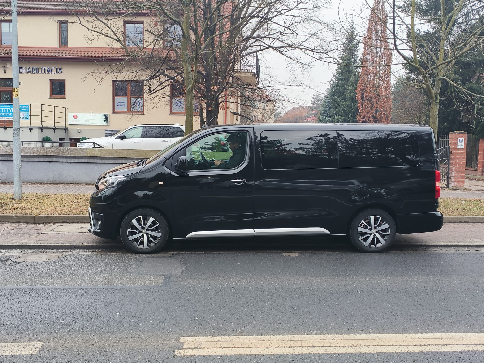 Czarny bus zaparkowany na ulicy, widoczny kierowca. W tle budynek z napisem 'Rehabilitacja' i drzewa. Auto z przyciemnianymi szybami.