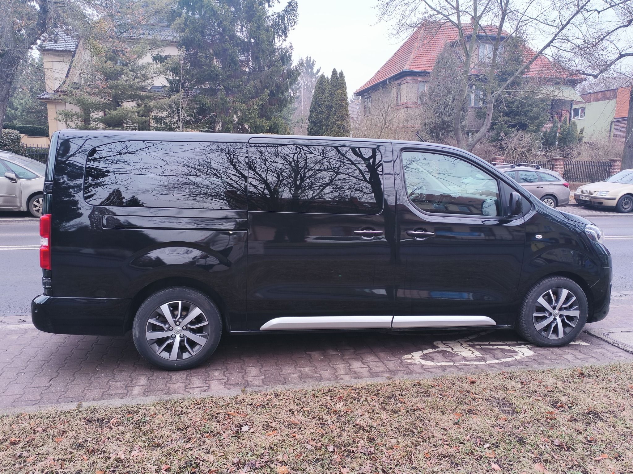 Czarny, duży bus zaparkowany na chodniku z kostki, z odbiciem drzew w szybach. W tle budynki mieszkalne i drzewa. Widok z boku, pochmurny dzień.
