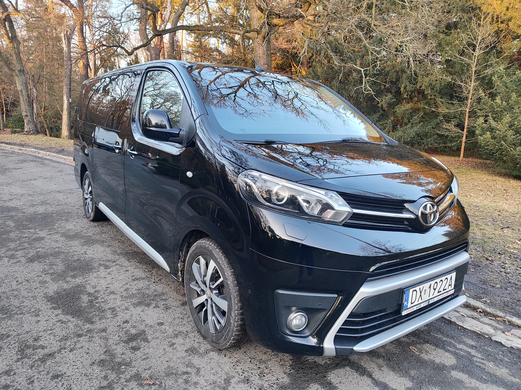 Czarny, lśniący bus Toyota Proace Verso z widoczną tablicą rejestracyjną DX 1922A, zaparkowany na tle drzew w parku, w słoneczny dzień.