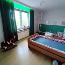 Pokój dziecięcy z drewnianym łóżkiem z plecionym ochraniaczem, zielonymi zasłonami z podświetleniem LED, drewnianym koniem na biegunach i drabinką gimnastyczną.