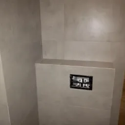 Szara, matowa ściana w łazience wyłożona płytkami z widoczną instalacją podtynkową do toalety, obudowana płytkami.